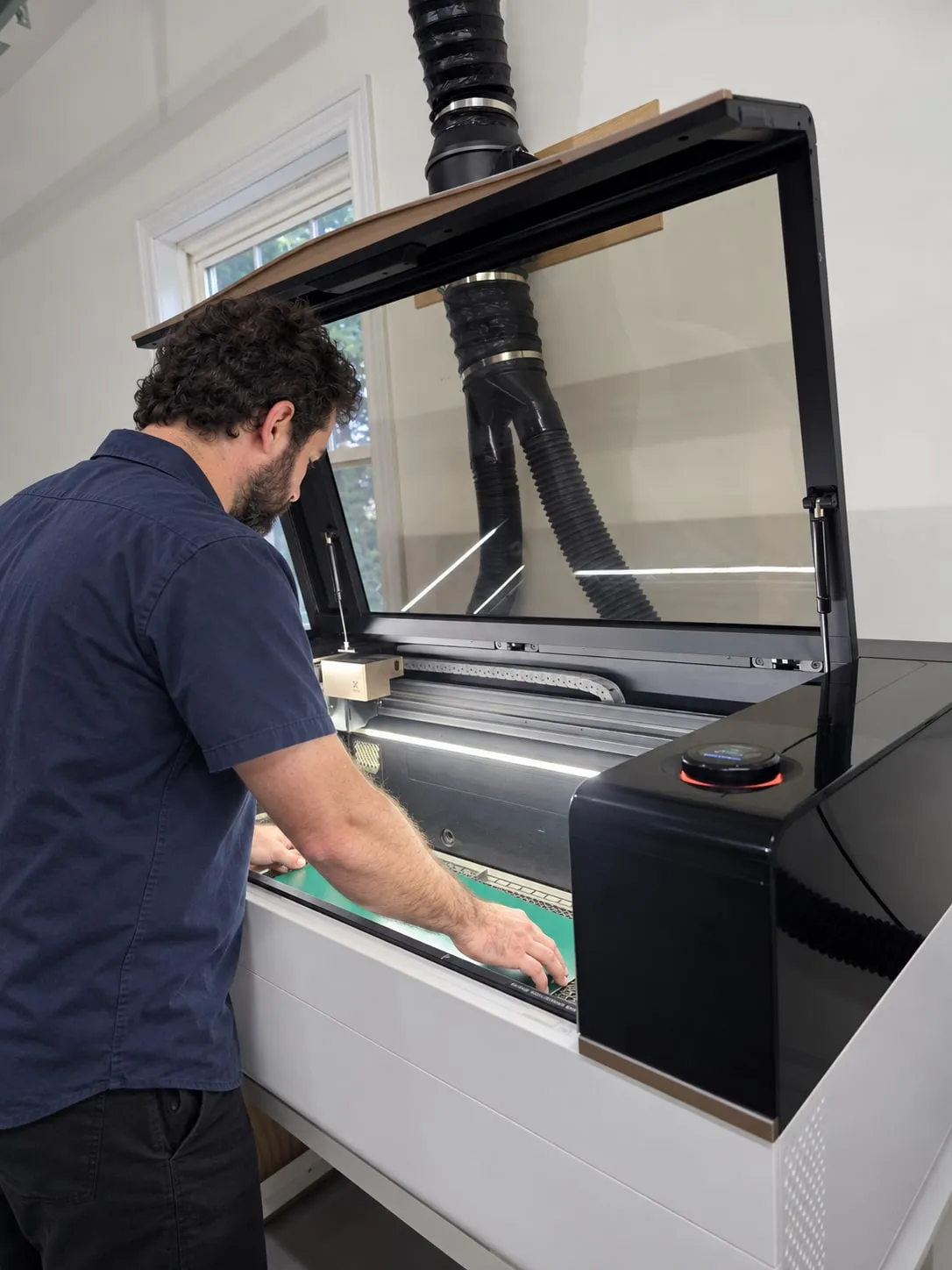 Someone loading material into the laser cutter, its lid raised and extraction ducting overhead