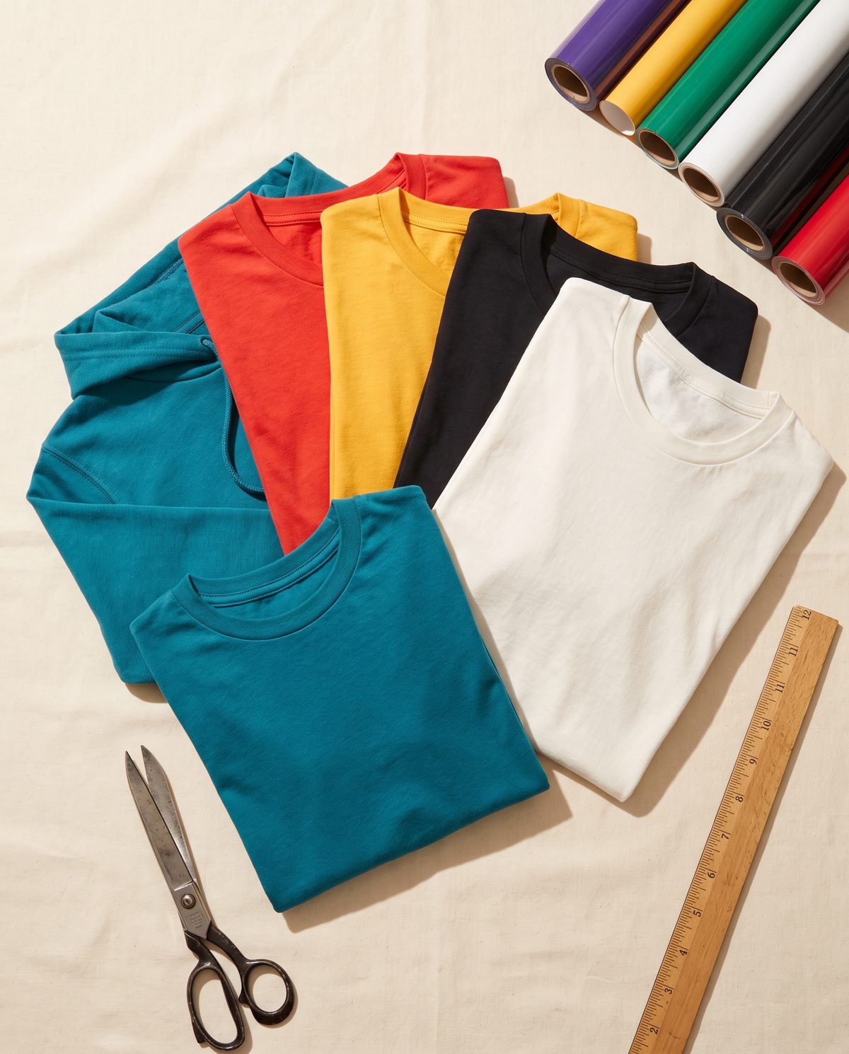 Folded blank tees and a hoodie in teal, red, gold and black beside vinyl and scissors